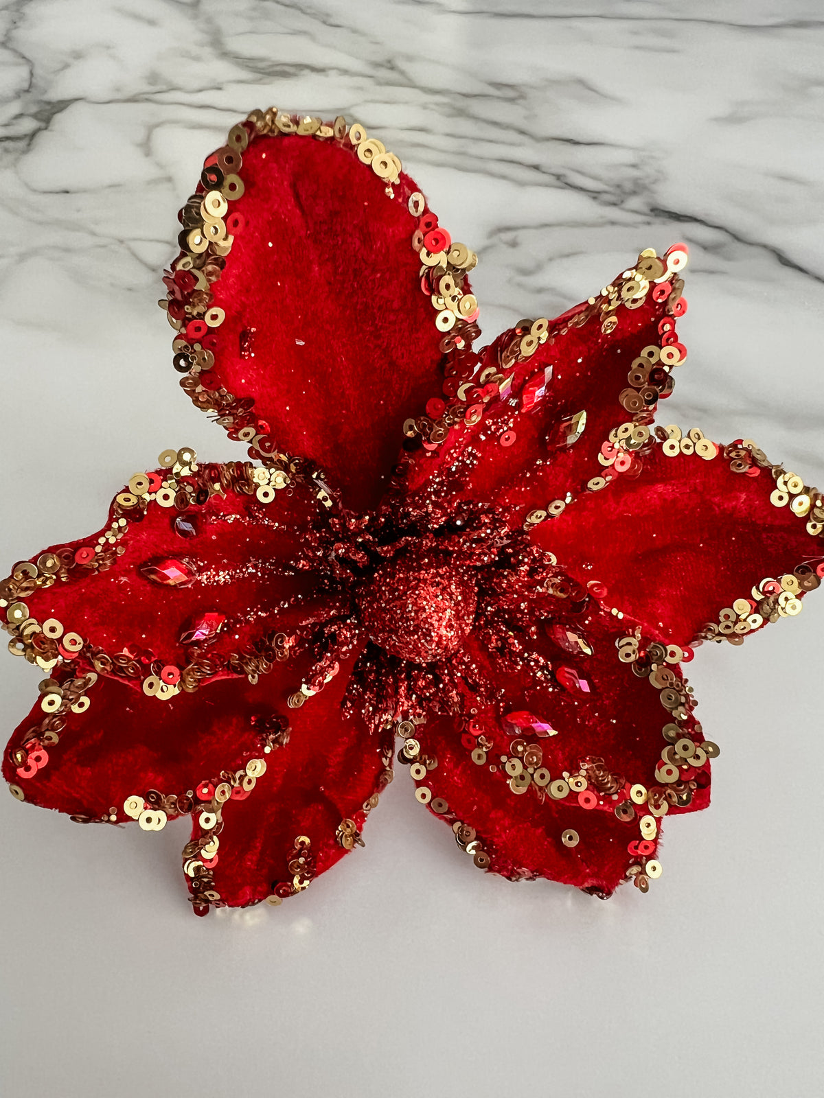 A red sequined artificial flower decoration with gold sequin trim, photographed on a white marble-patterned surface.