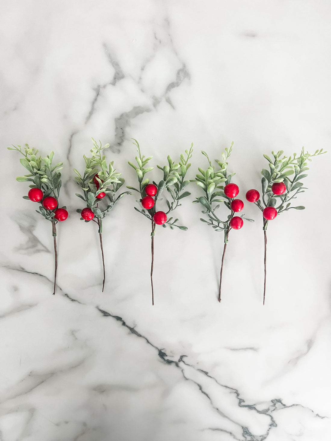 Five artificial greenery sticks with red berries on a marble surface