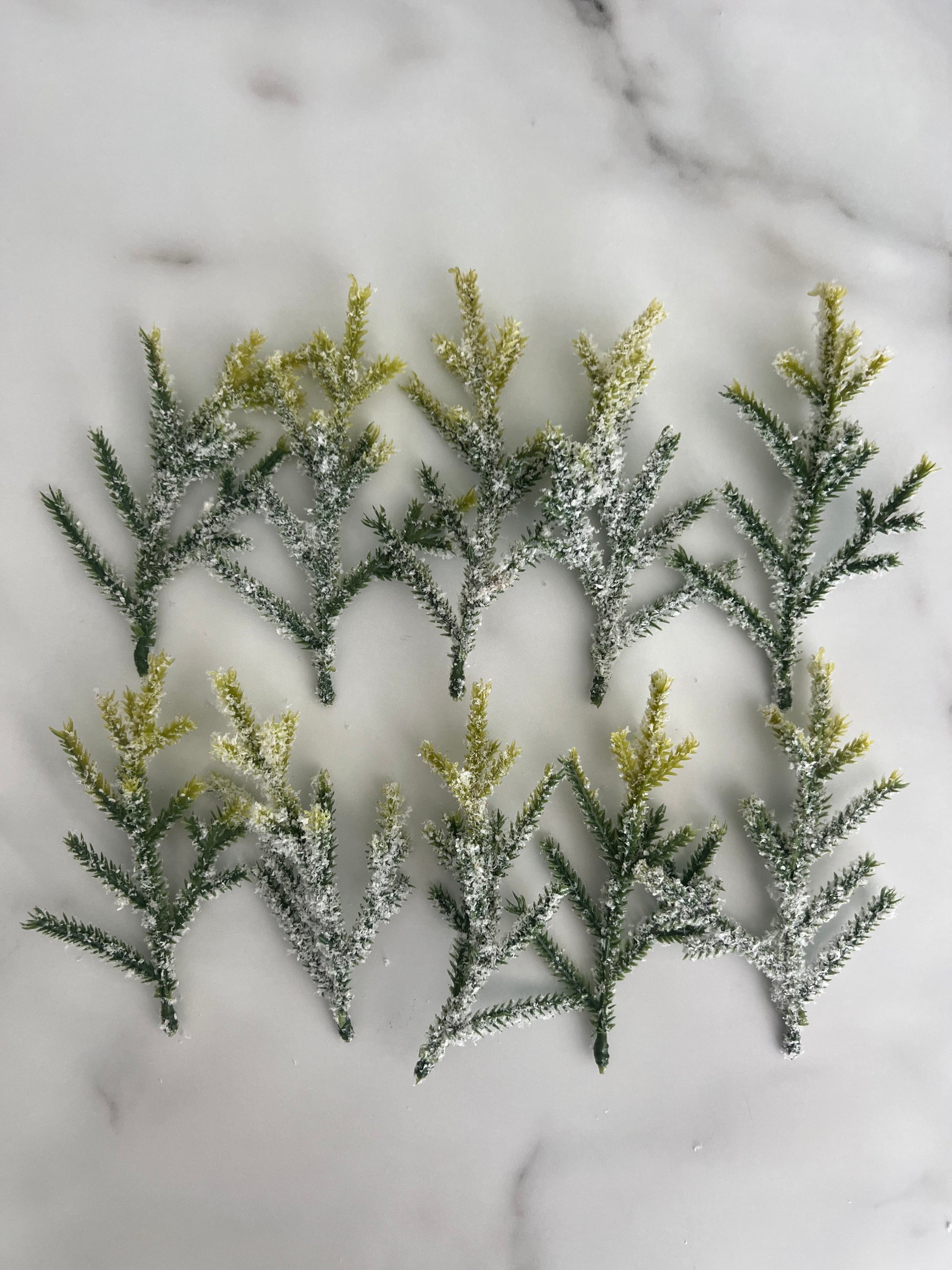 Ten artificial frosted pine branches with green needles and yellow-tipped needles, arranged on a white marble surface.