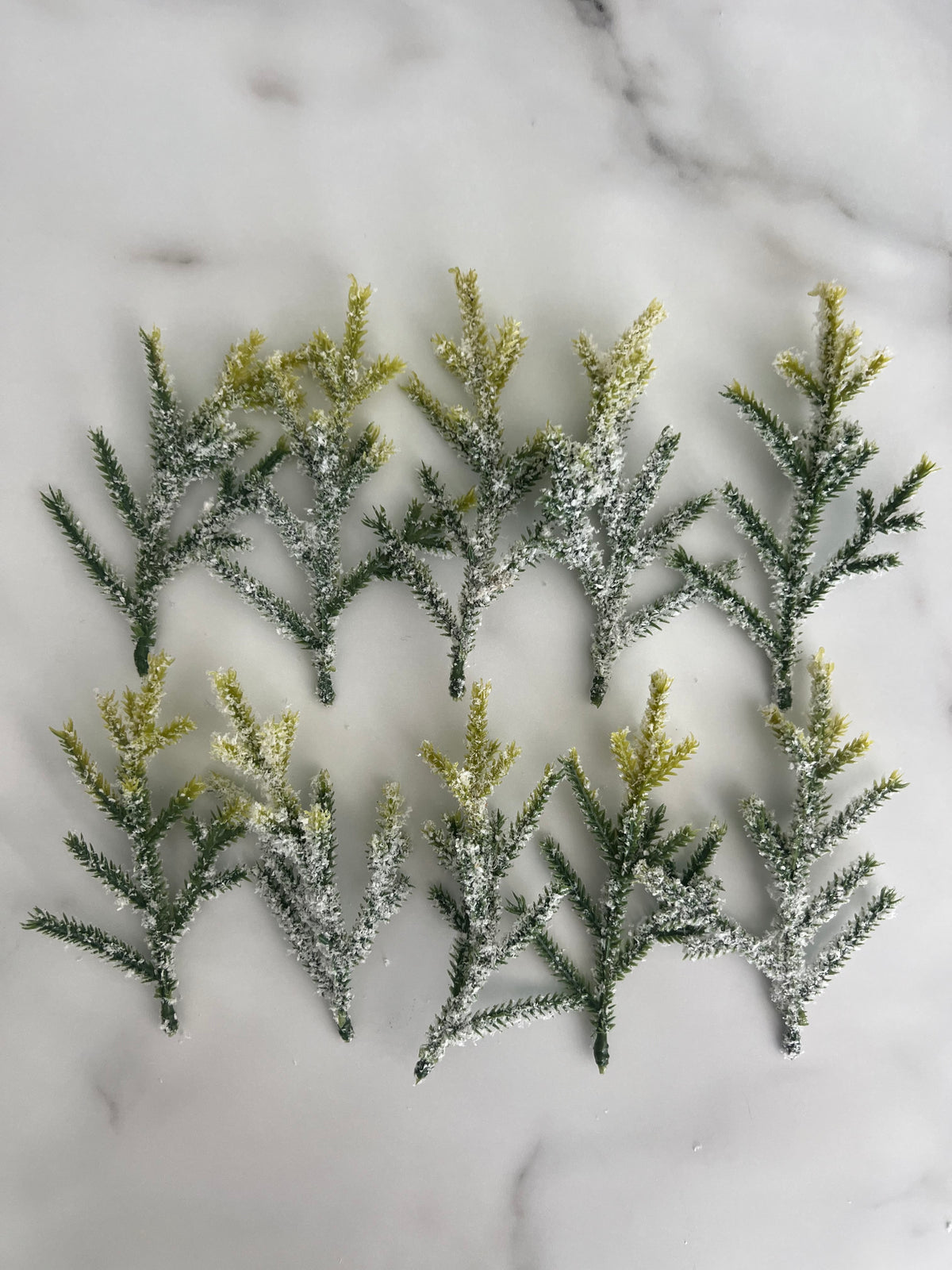 Ten artificial frosted pine branches with green needles and yellow-tipped needles, arranged on a white marble surface.