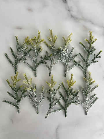 Ten artificial frosted pine branches with green needles and yellow-tipped needles, arranged on a white marble surface.