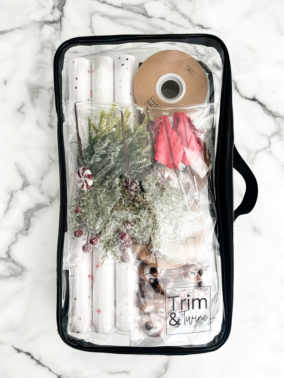 A clear plastic carrying case with a black zipper and handle, filled with holiday wrapping supplies including rolls of  wrapping paper and wrapping items. The case is photographed from above on a white marble surface.