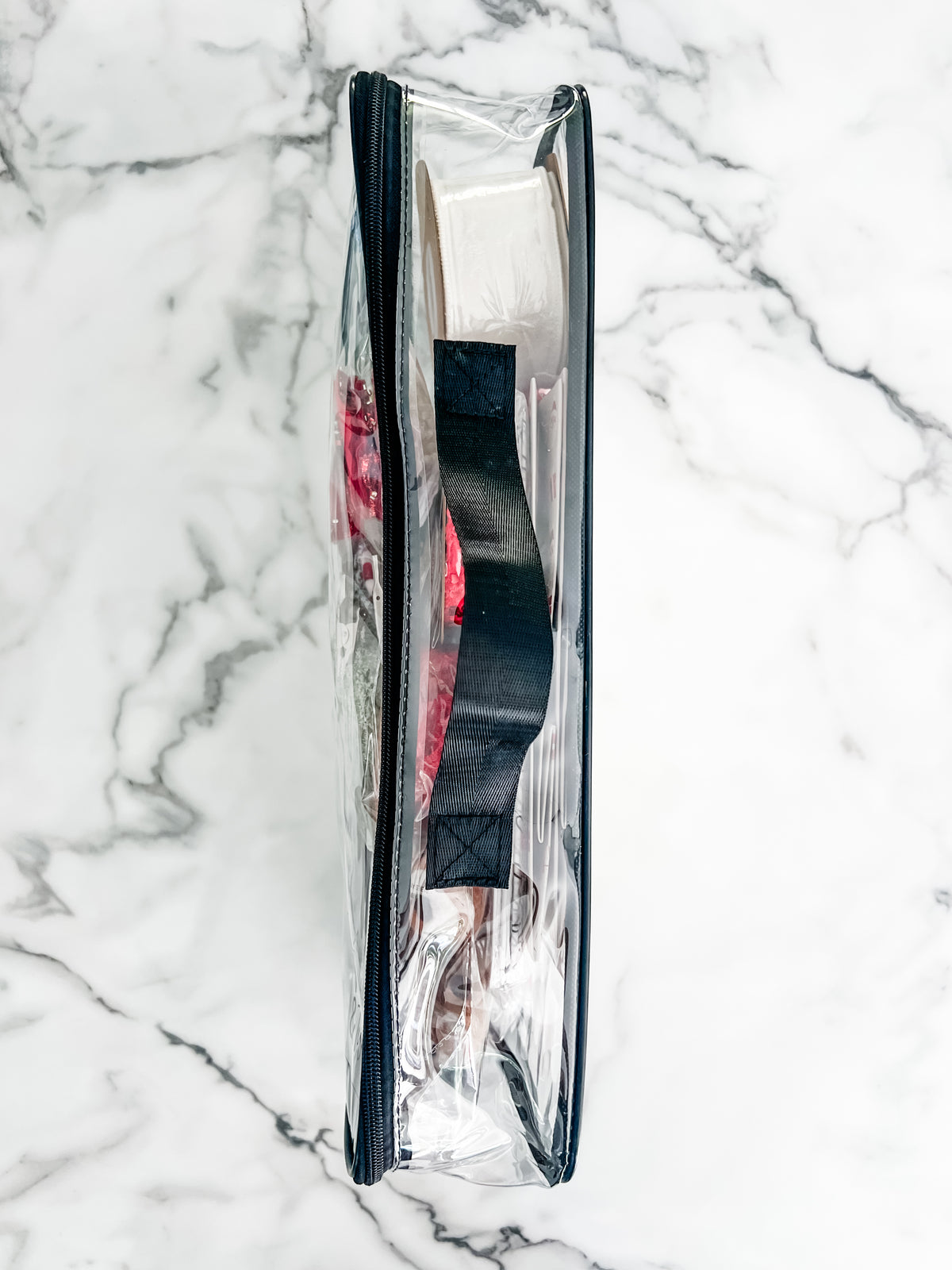 A clear plastic carrying case with a black zipper and handle, filled with holiday wrapping supplies including rolls of  wrapping paper and wrapping items. The case is photographed from above on a white marble surface.