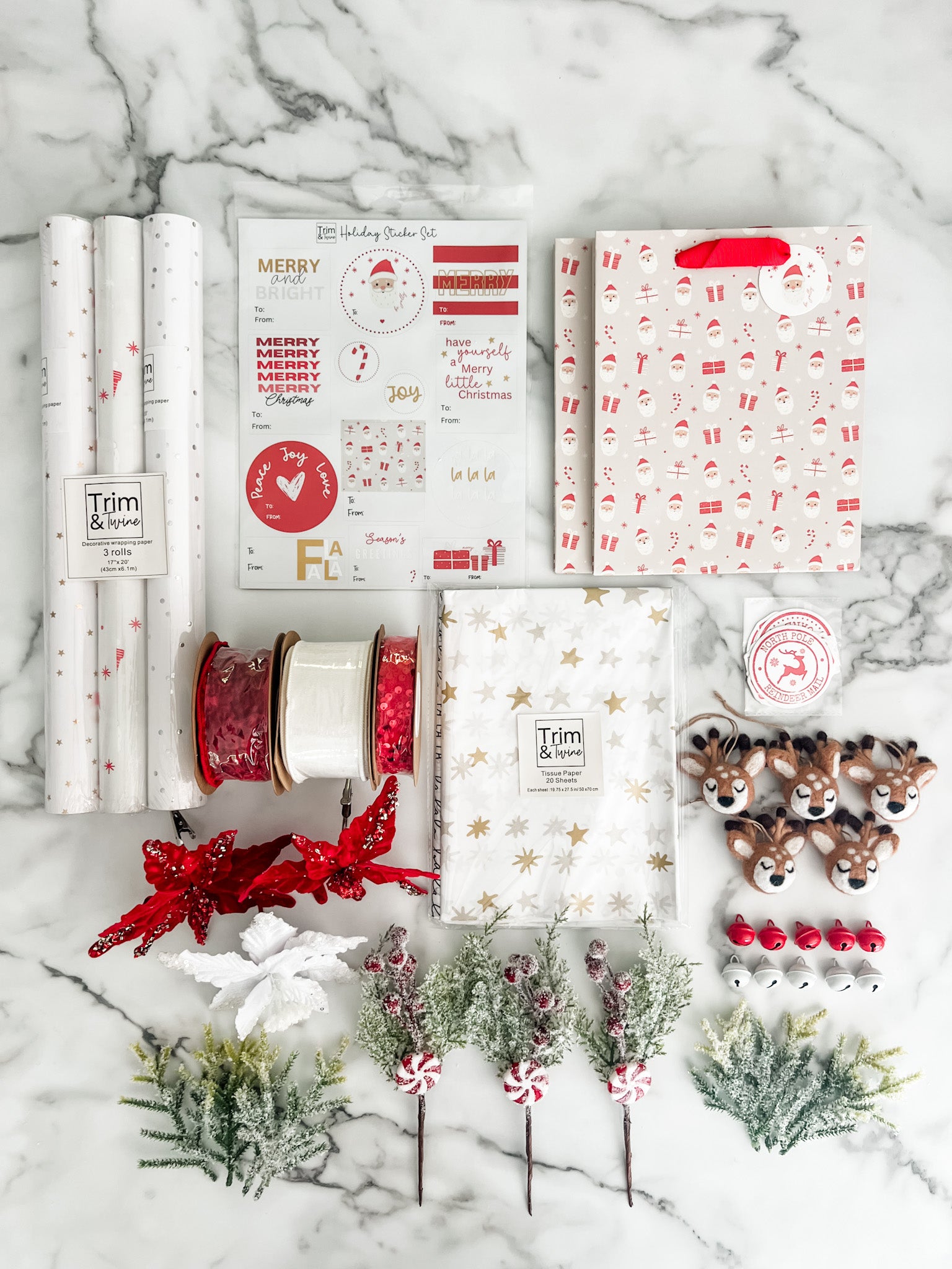 A flat-lay arrangement of Christmas wrapping supplies and decorations on a white marble surface, including patterned wrapping paper rolls, gift bags, ribbon spools, stickers, reindeer figurines, and small greenery accents.