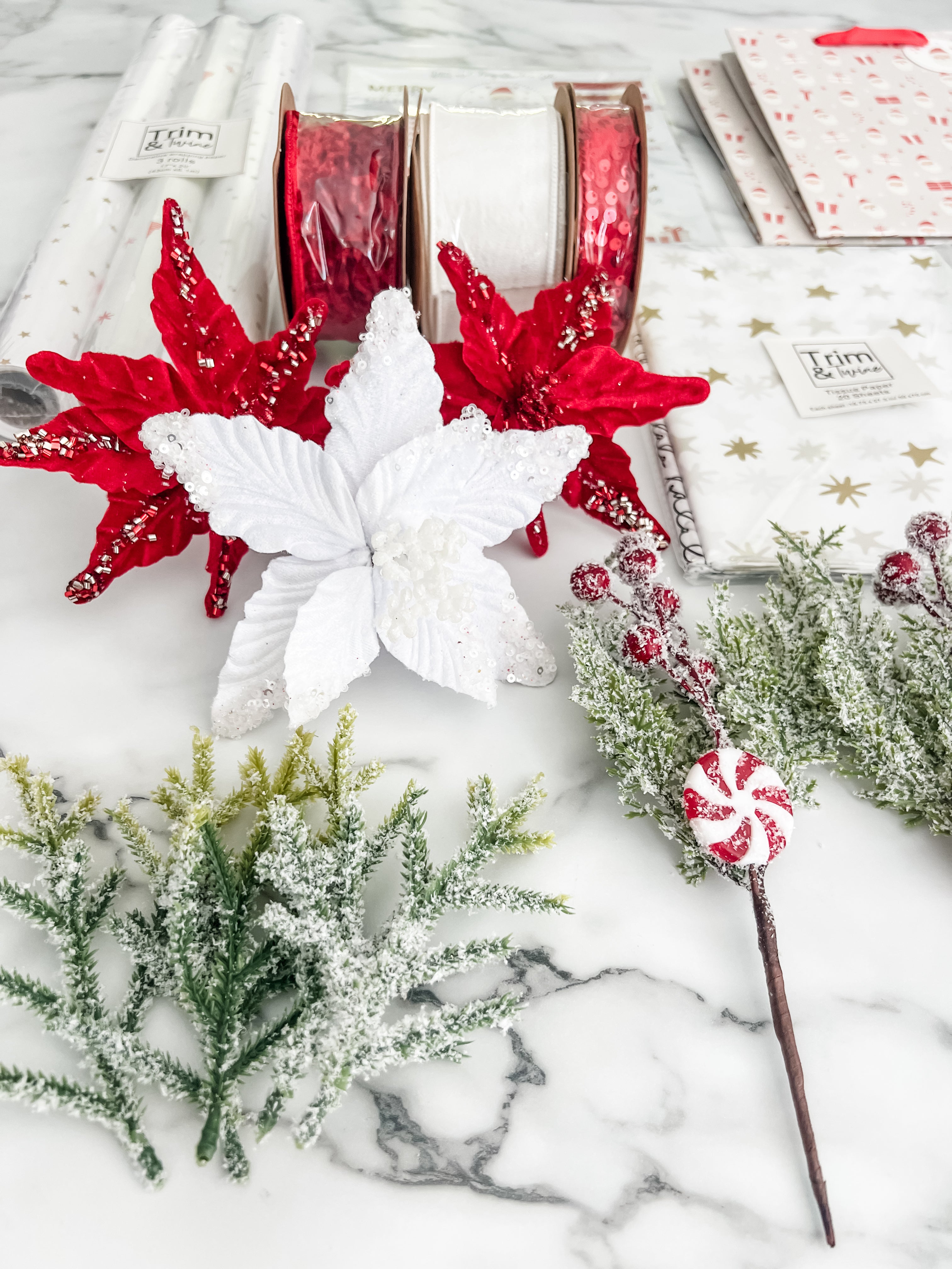 A styled flat-lay of Christmas-themed stationery and decor on a white marble surface, including red and white ribbon spools, white and red poinsettias, frosted greenery, and a peppermint stick.