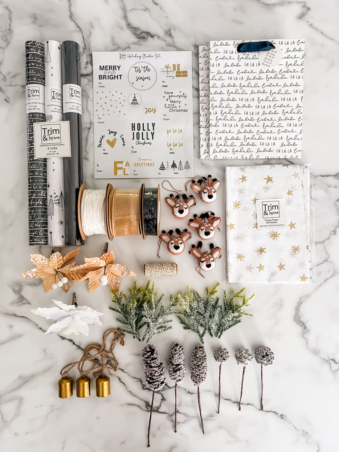 A styled flat-lay of a holiday-themed crafting kit on a white marble background, featuring wrapping paper rolls, stamps, stickers, glitter, pinecones, faux greenery, and a wrapped gift box.
