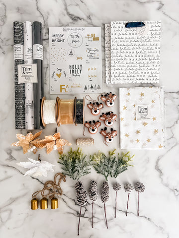A styled flat-lay of a holiday-themed crafting kit on a white marble background, featuring wrapping paper rolls, stamps, stickers, glitter, pinecones, faux greenery, and a wrapped gift box.