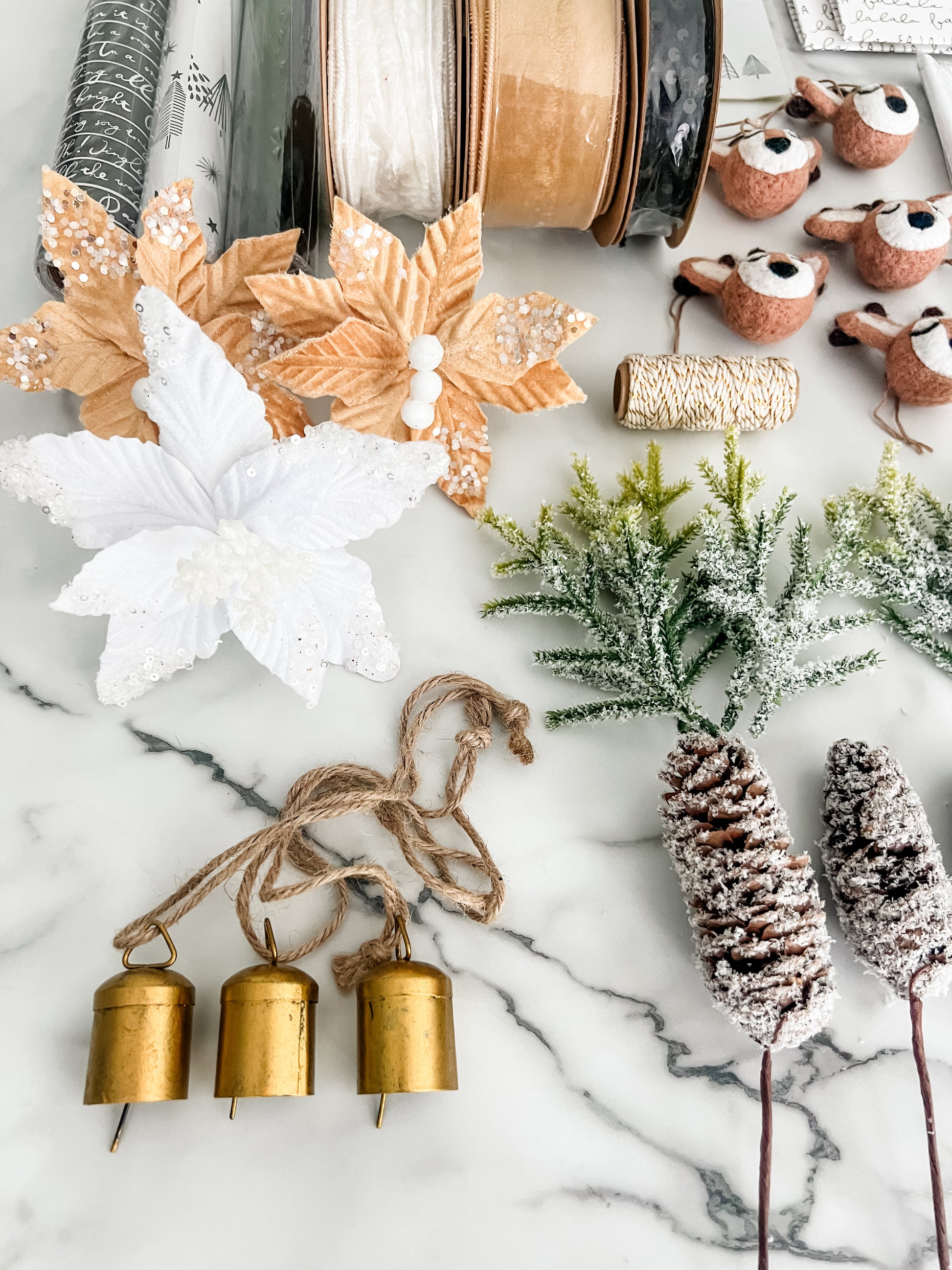 A styled flat-lay of Christmas craft supplies and decorations on a white marble background, including faux poinsettias in white and gold, ribbon spools, deer embellishments, twine, green frosted branches, pinecones, and three small gold bells.