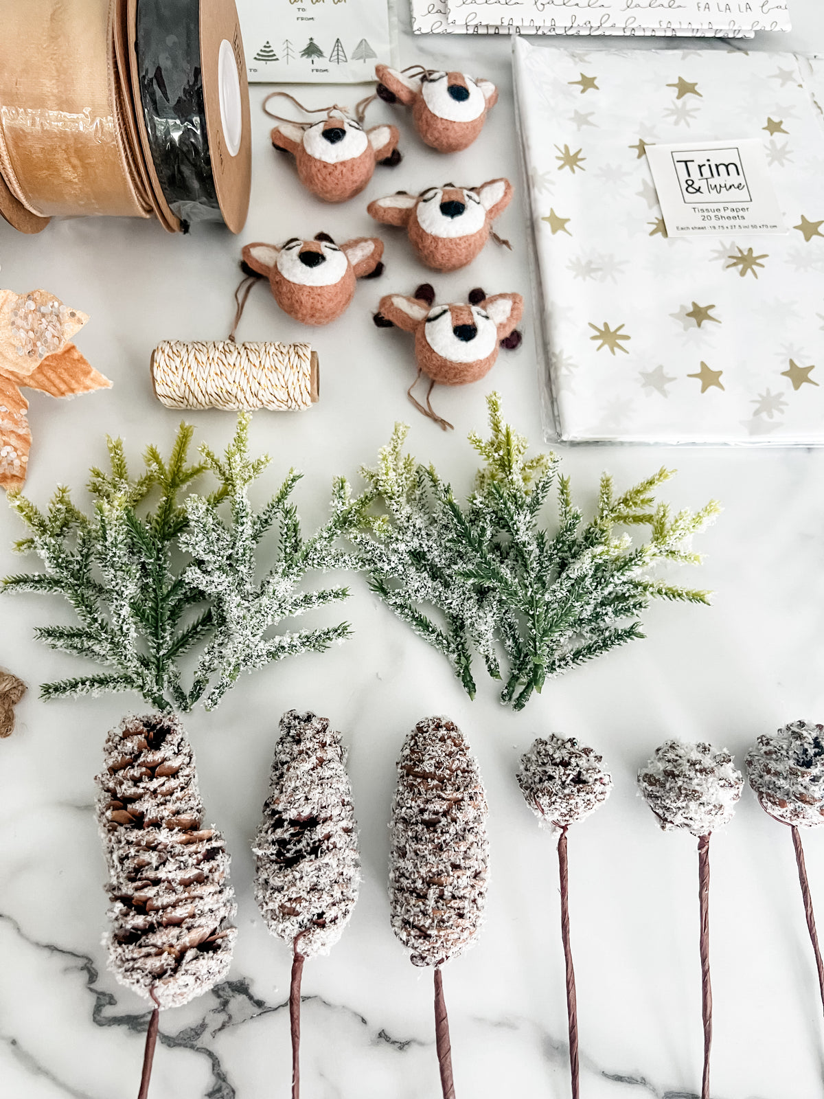 A styled flat-lay of Christmas decoration supplies on a white marble surface, including faux pine branches, pinecone picks, reindeer head ornaments, ribbon spools, and a gift bag with gold stars and the words 'Trim & Tinsel'.