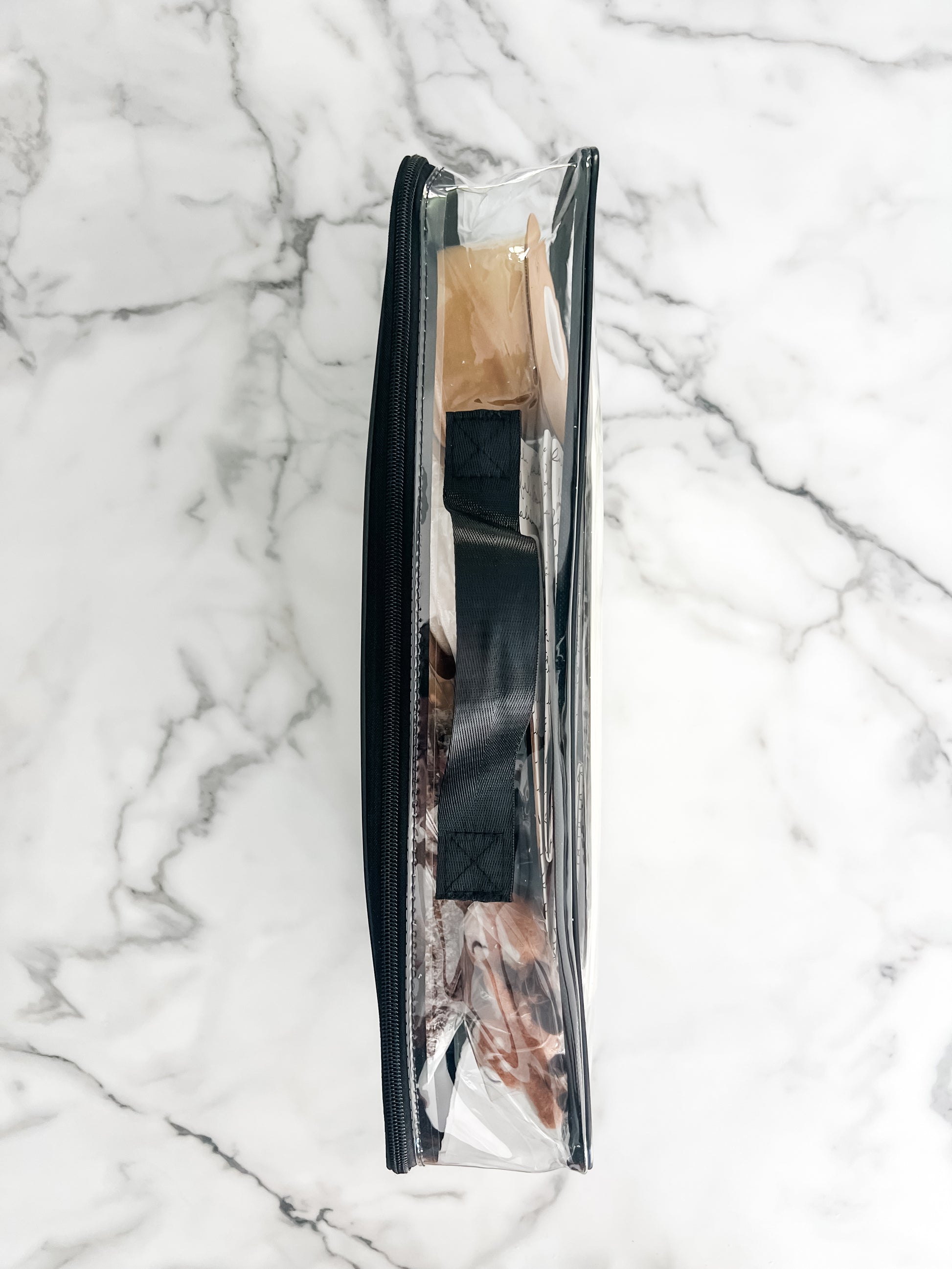 A clear zippered storage bag with a black handle and items inside, laid flat on a white marble surface. 
