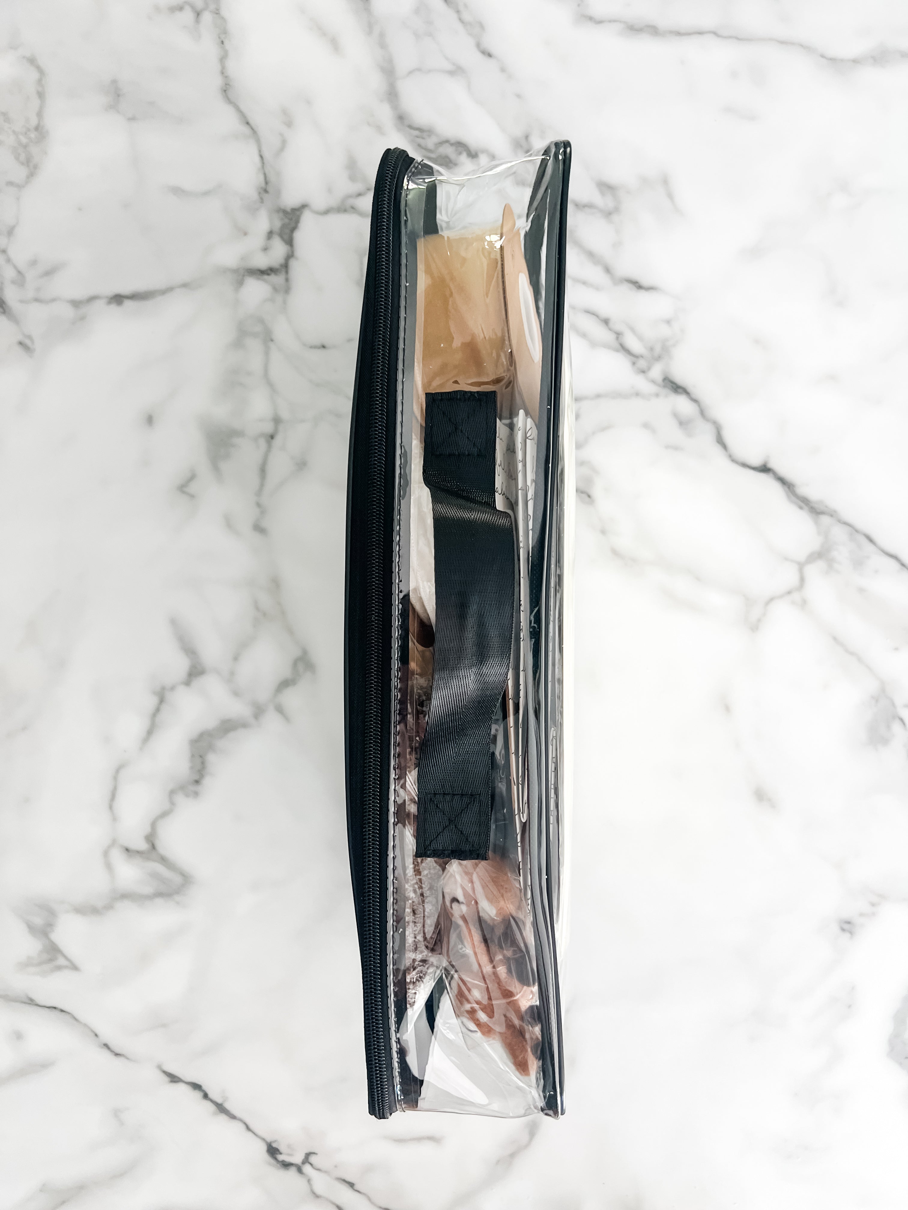 A clear zippered storage bag with a black handle and items inside, laid flat on a white marble surface. 