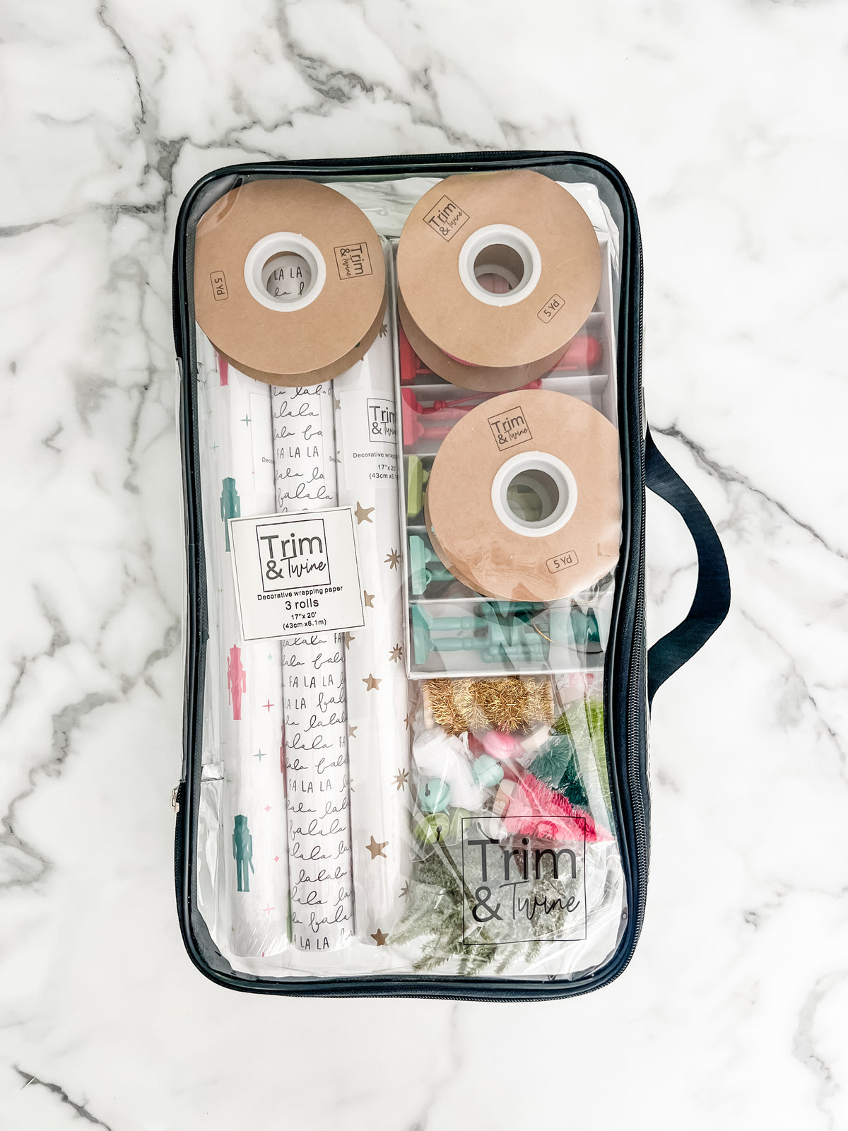 A clear zippered storage bag with a black handle, laid flat on a white marble surface. Inside are several rolls of wrapping paper and other wrapping items with visible Trim & Twine branding.