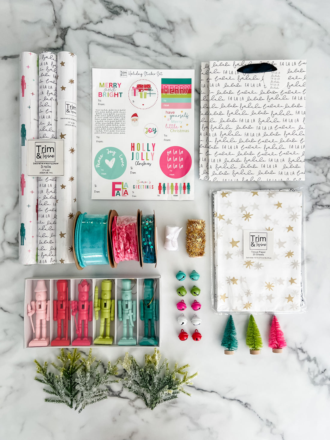 A flat lay of a Christmas-themed crafting kit on a white marble background, including patterned wrapping paper rolls, ribbon spools, small ornaments, Christmas trees, and printed cards and stickers.