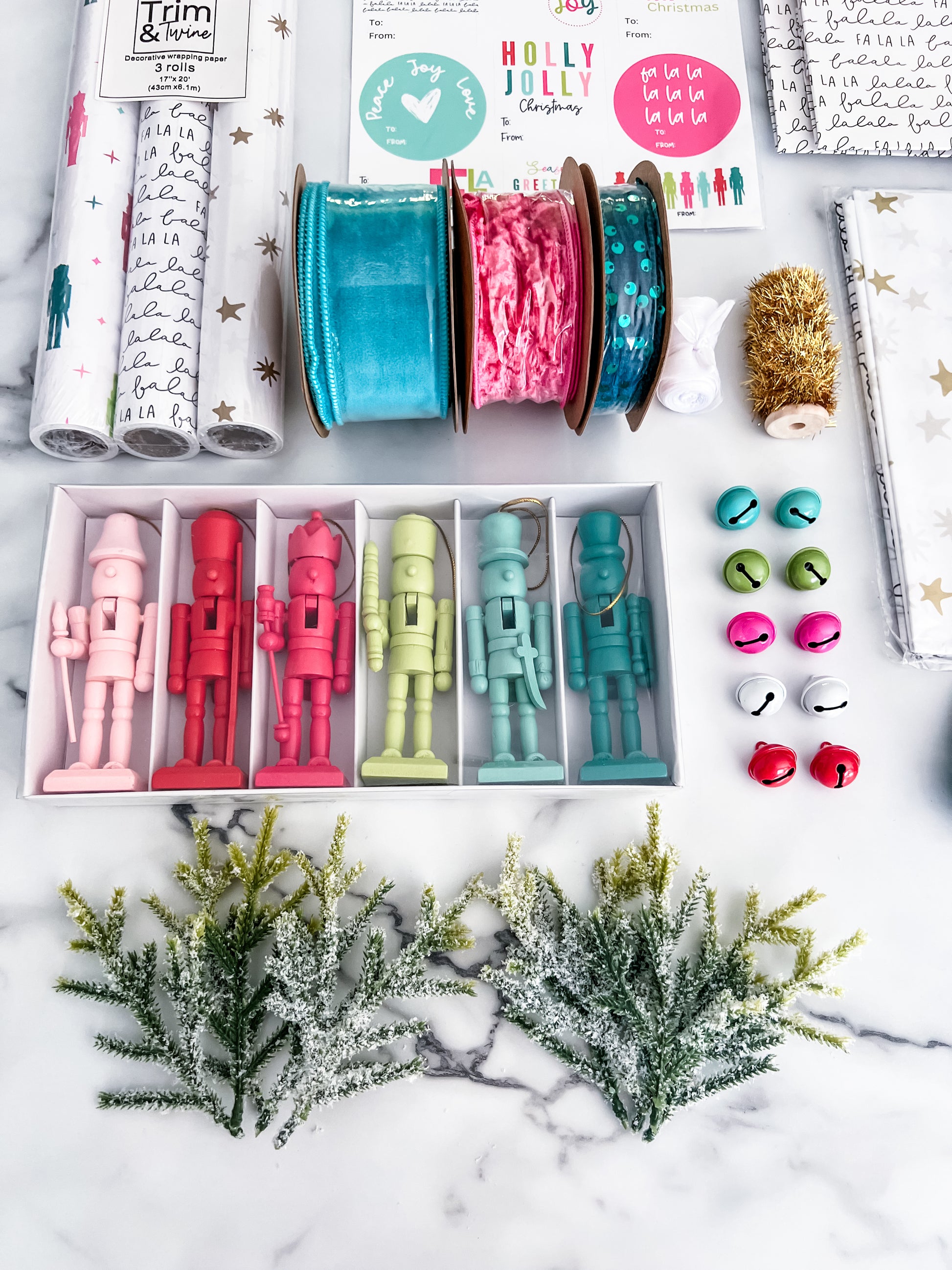 A flat lay of a Christmas-themed crafting kit on a white marble background, including patterned wrapping paper rolls, ribbon spools, small ornaments, Christmas trees, and printed cards and stickers.