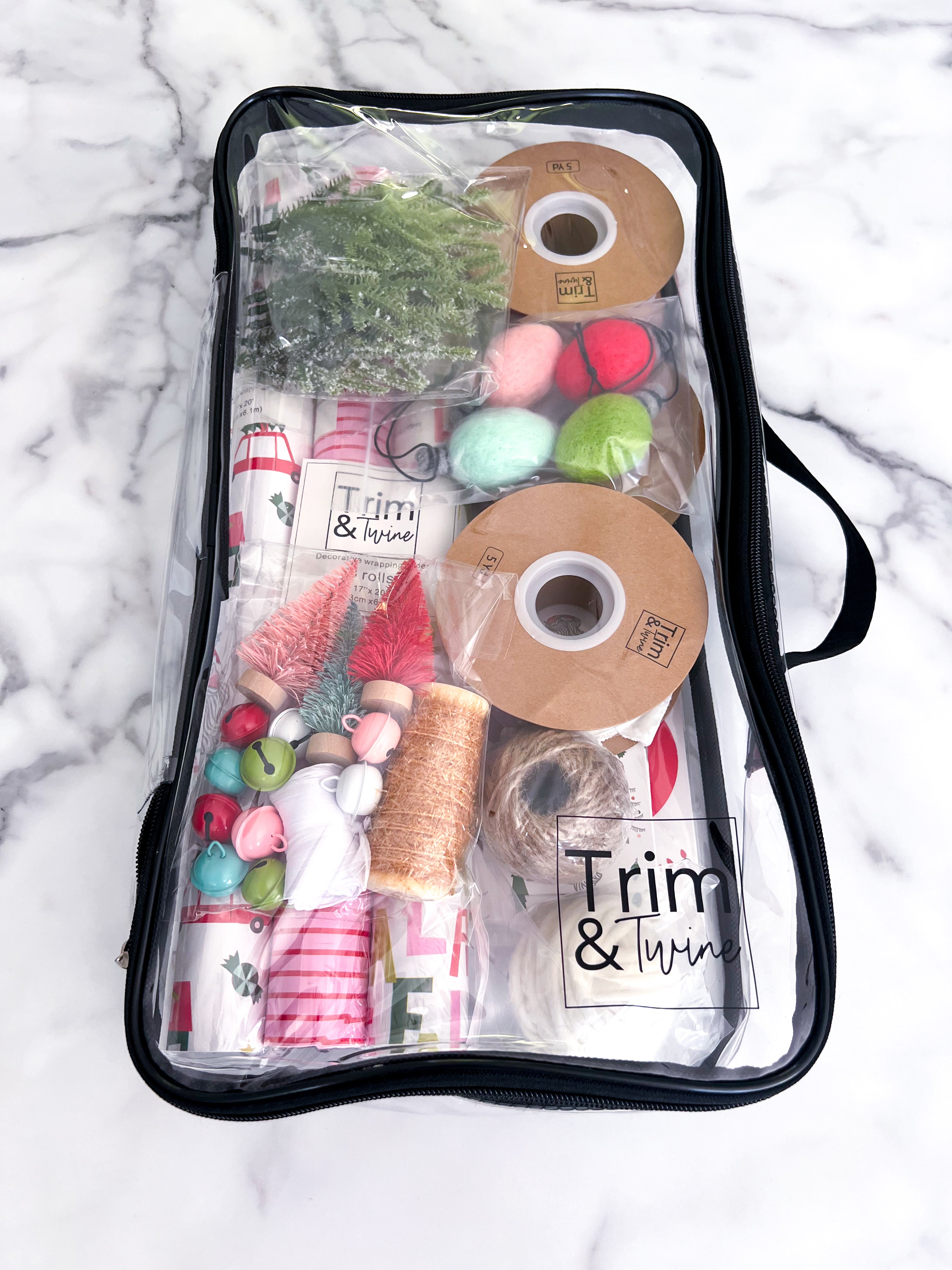 A clear zippered storage bag with a black zipper and handle, filled with various items including spools of tinsel, ribbon, bells, and small Christmas trees, photographed from above on a white marble surface. The brand name 'Trim & Twine' is visible on a label and on some of the included items.