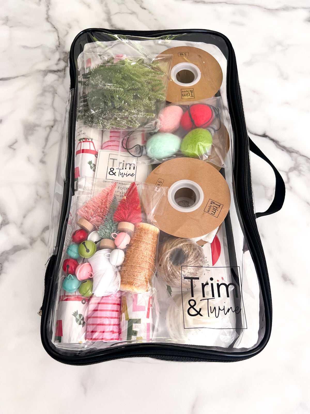 A clear zippered storage bag with a black zipper and handle, filled with various items including spools of tinsel, ribbon, bells, and small Christmas trees, photographed from above on a white marble surface. The brand name 'Trim & Twine' is visible on a label and on some of the included items.
