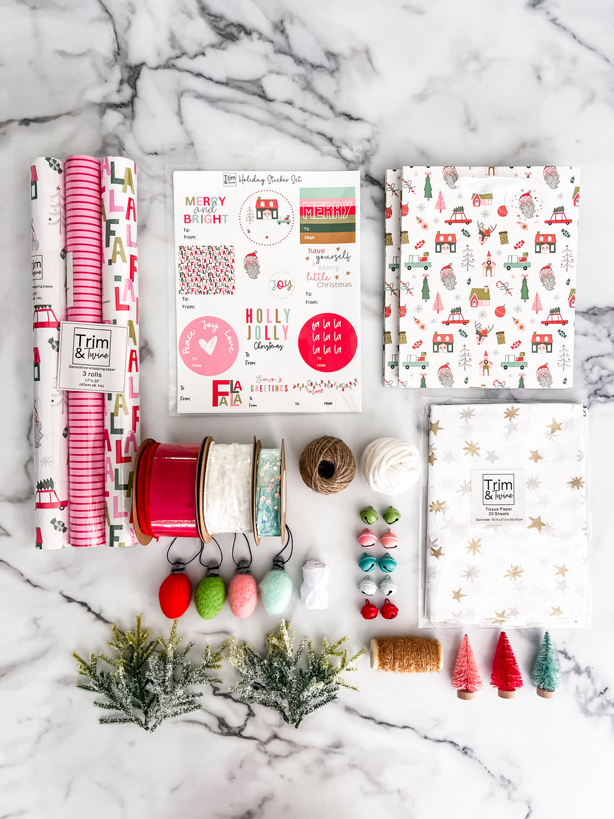 A flat-lay arrangement of Christmas-themed crafting supplies on a white marble background, including patterned wrapping paper rolls, a sticker sheet, gift tags, ribbon spools, bells, tinsel, small pine branches, and mini Christmas trees.