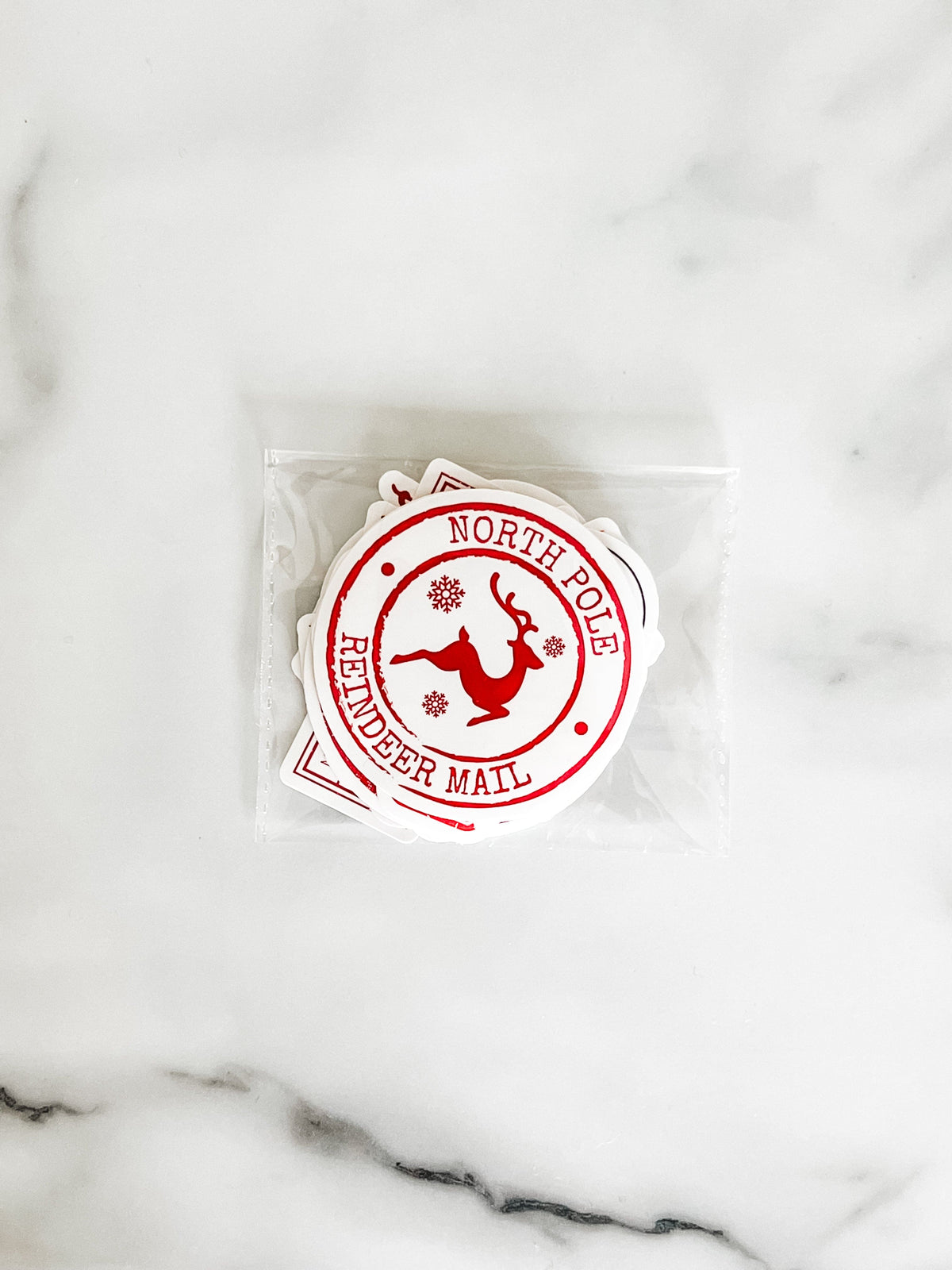 A set of white die-cut stickers featuring a red circular North Pole Reindeer Mail stamp design, sealed in a clear plastic bag on a white marble surface.