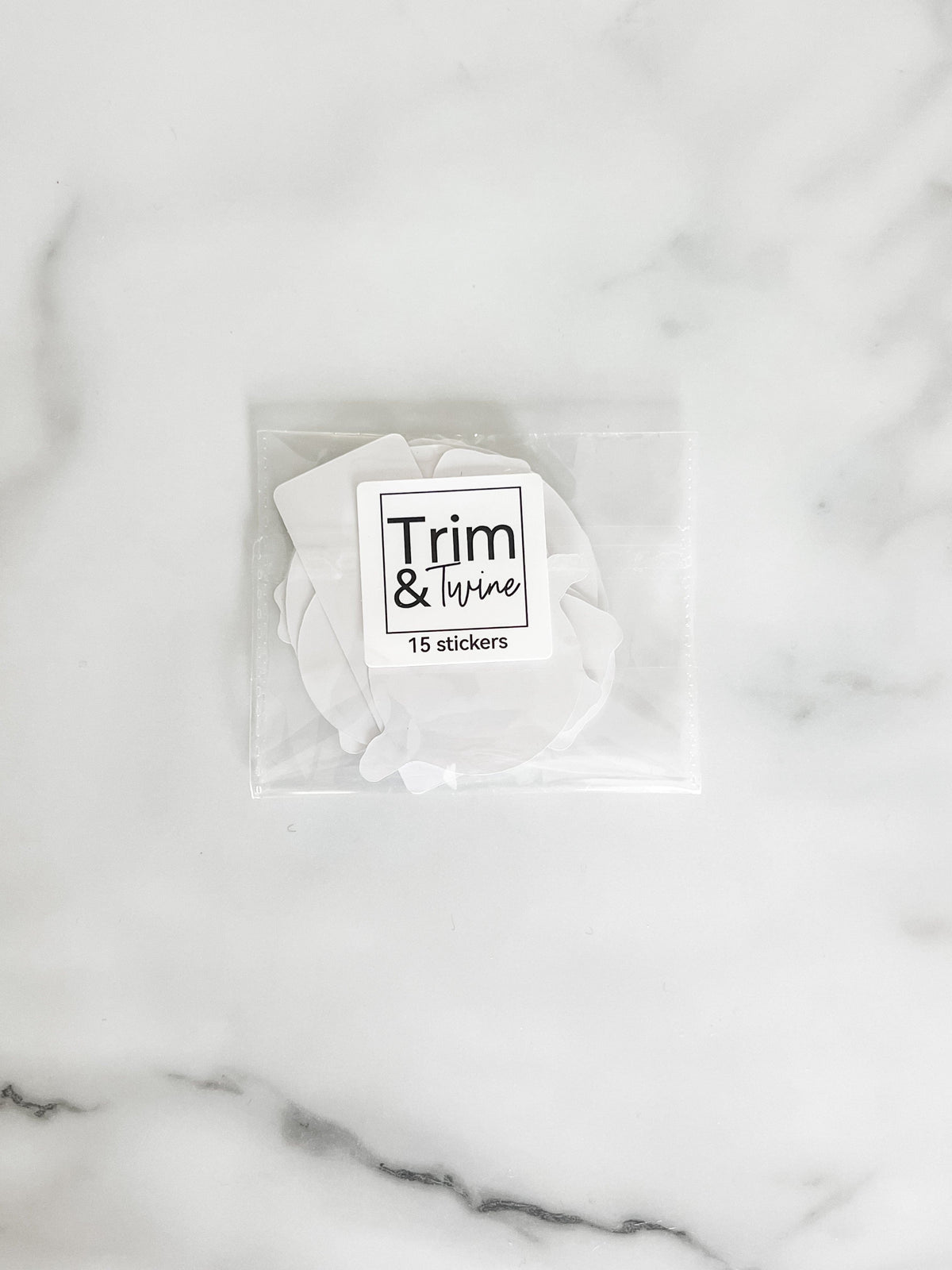 A small clear plastic bag containing white circular stickers with the text 'Trim & tidy 15 stickers' on a white marble surface.