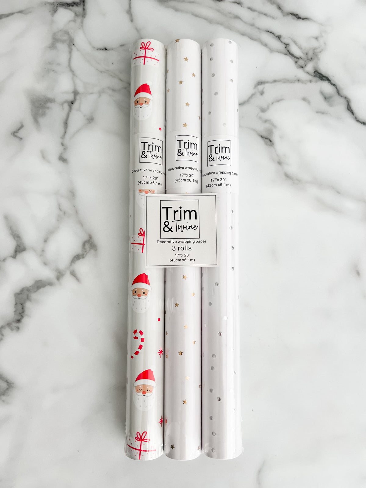 Three rolls of wrapping paper laid out next to each other, with one roll featuring a santa pattern, another with gold stars, and the last one with metallic silver dots. 