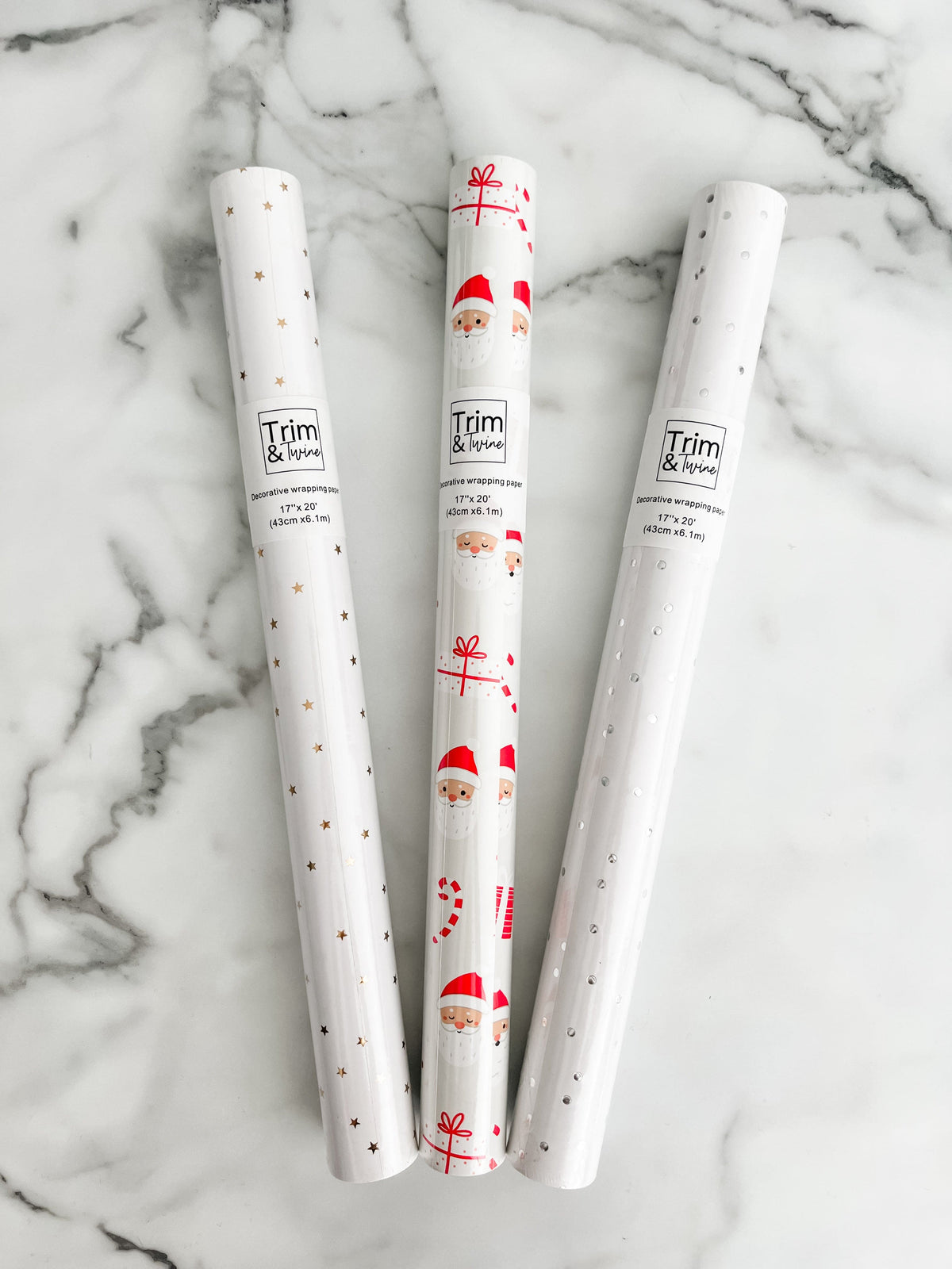 Three rolls of wrapping paper with Santa Claus design, metallic gold stars, and metallic silver dots, placed on a marble surface.