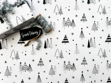 Wrapping paper with a Christmas tree and star pattern in black and white colors, laying on a white surface with a tag and silver tinsel.