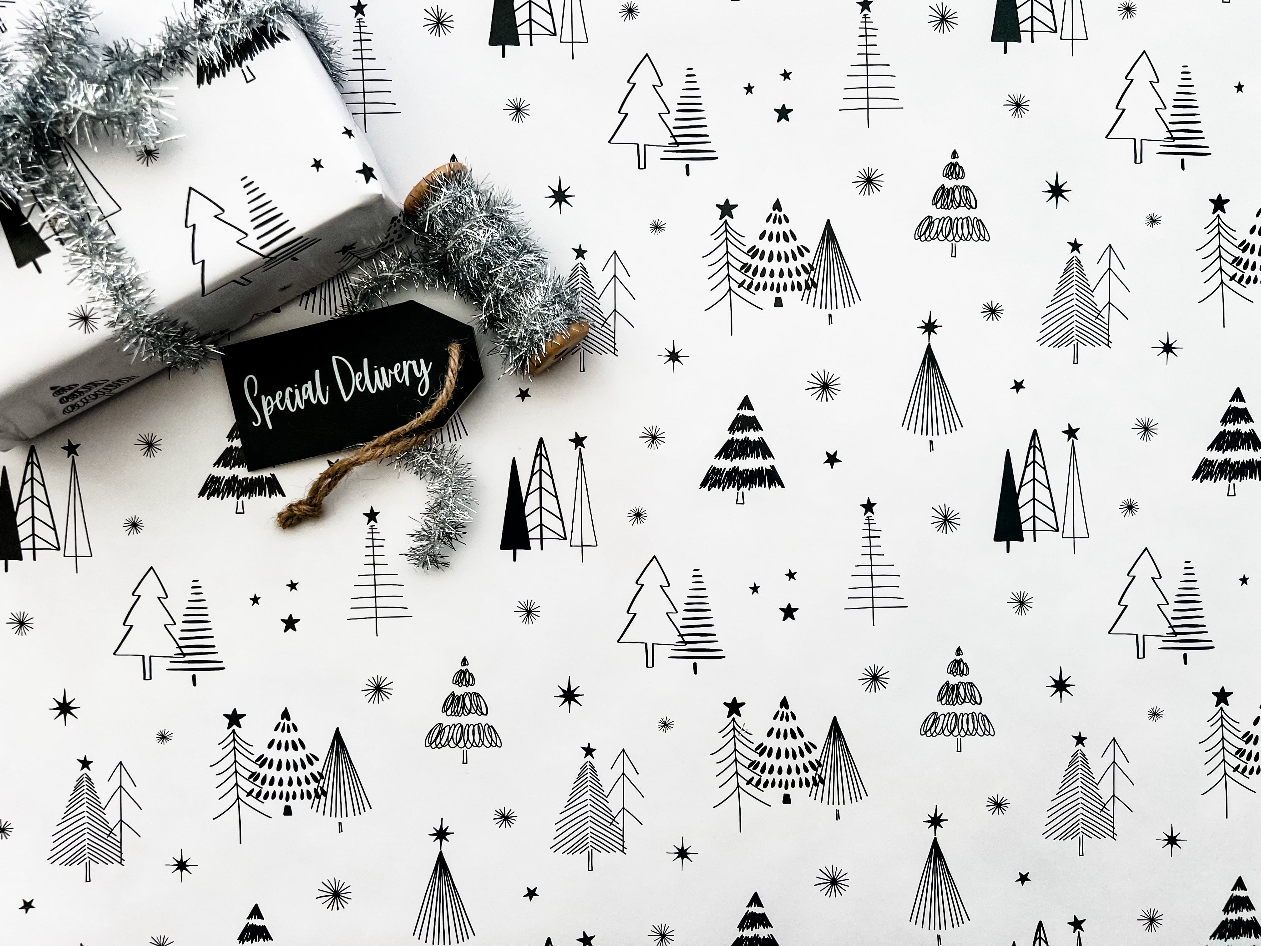 Wrapping paper with a Christmas tree and star pattern in black and white colors, laying on a white surface with a tag and silver tinsel.