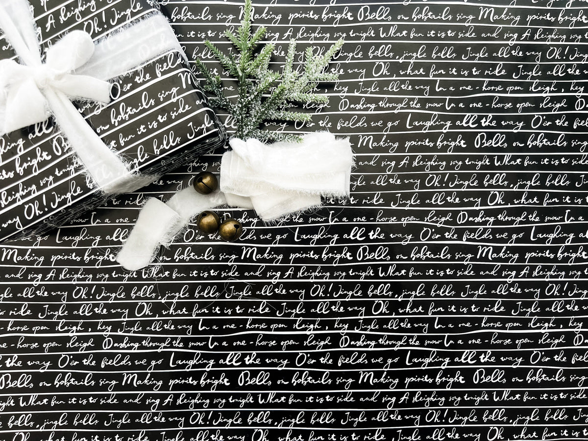 A roll of wrapping paper with 'Jingle Bells' lyrics printed in white text on a black background, displayed alongside Christmas decorations.