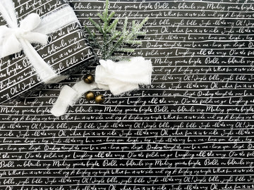 A roll of wrapping paper with 'Jingle Bells' lyrics printed in white text on a black background, displayed alongside Christmas decorations.