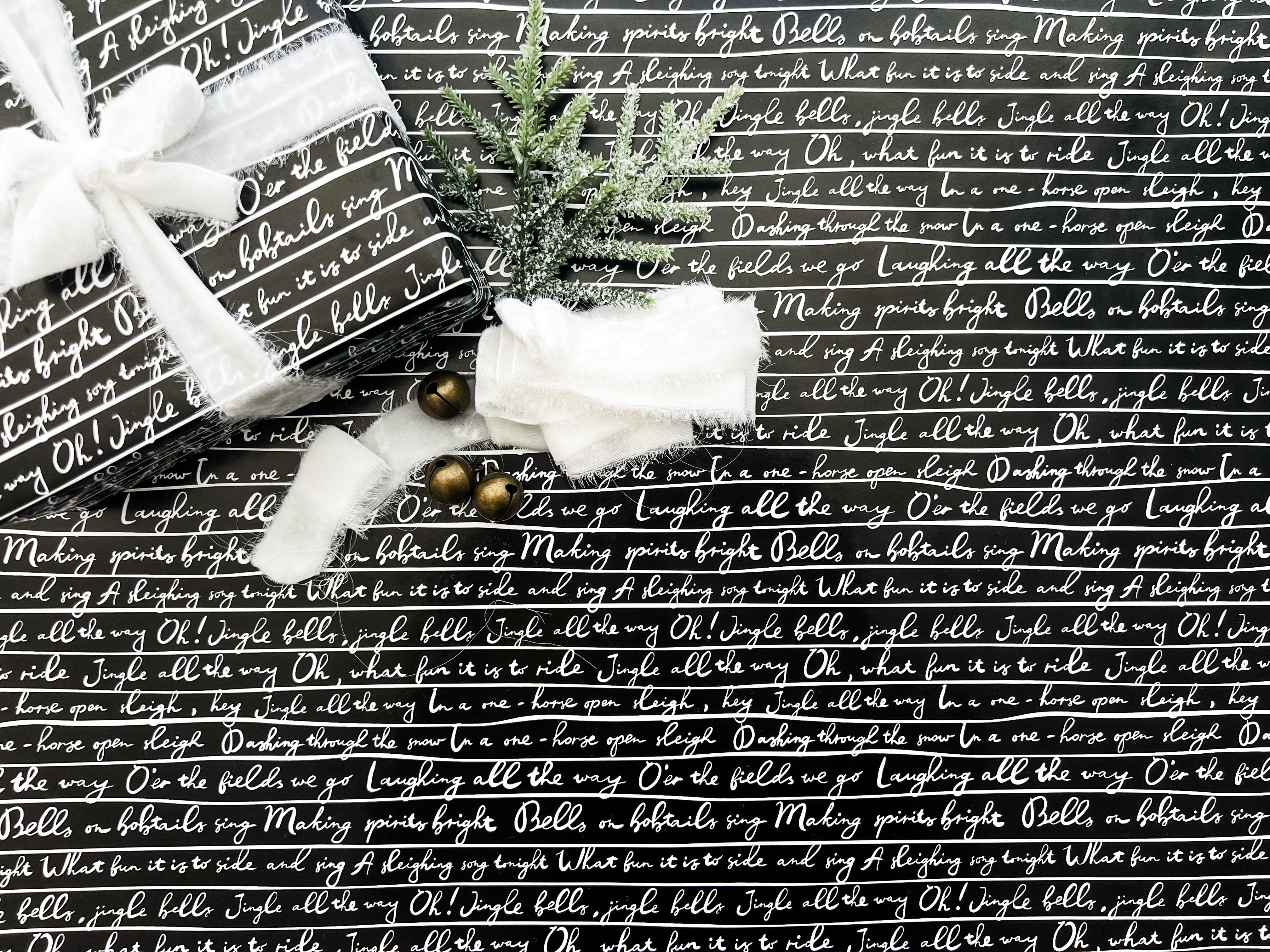 A roll of wrapping paper with 'Jingle Bells' lyrics printed in white text on a black background, displayed alongside Christmas decorations.