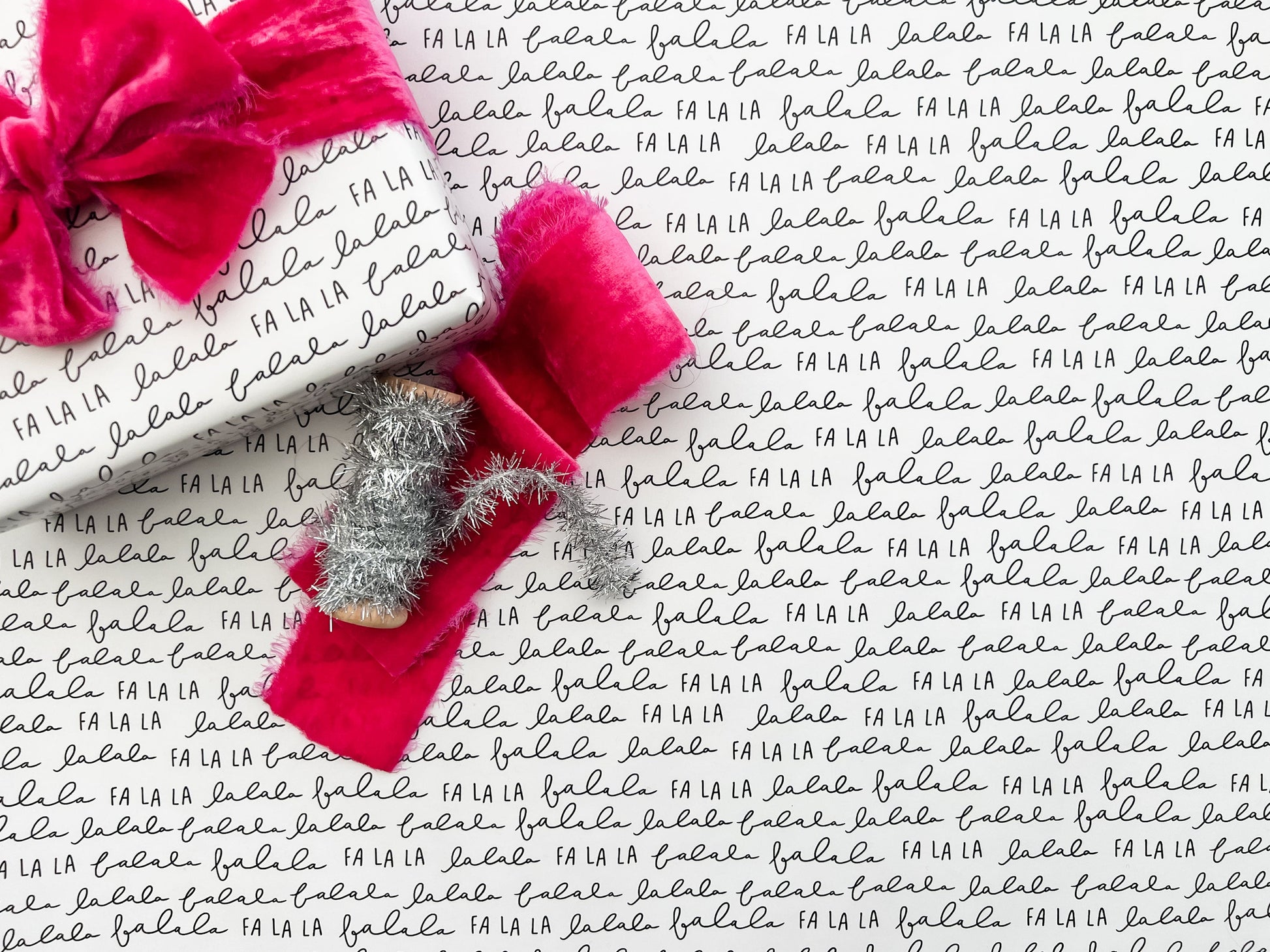Flat lay of white wrapping paper with repeating script pattern of fa la la.  Shown with a hot pink velvet bow and silver tinsel.