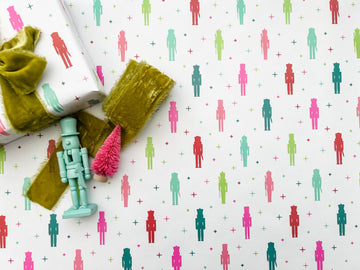 A laid out roll of wrapping paper with a colorful nutcracker pattern featuring nutcracker figures, displayed alongside a green ribbon and a decorative nutcracker.