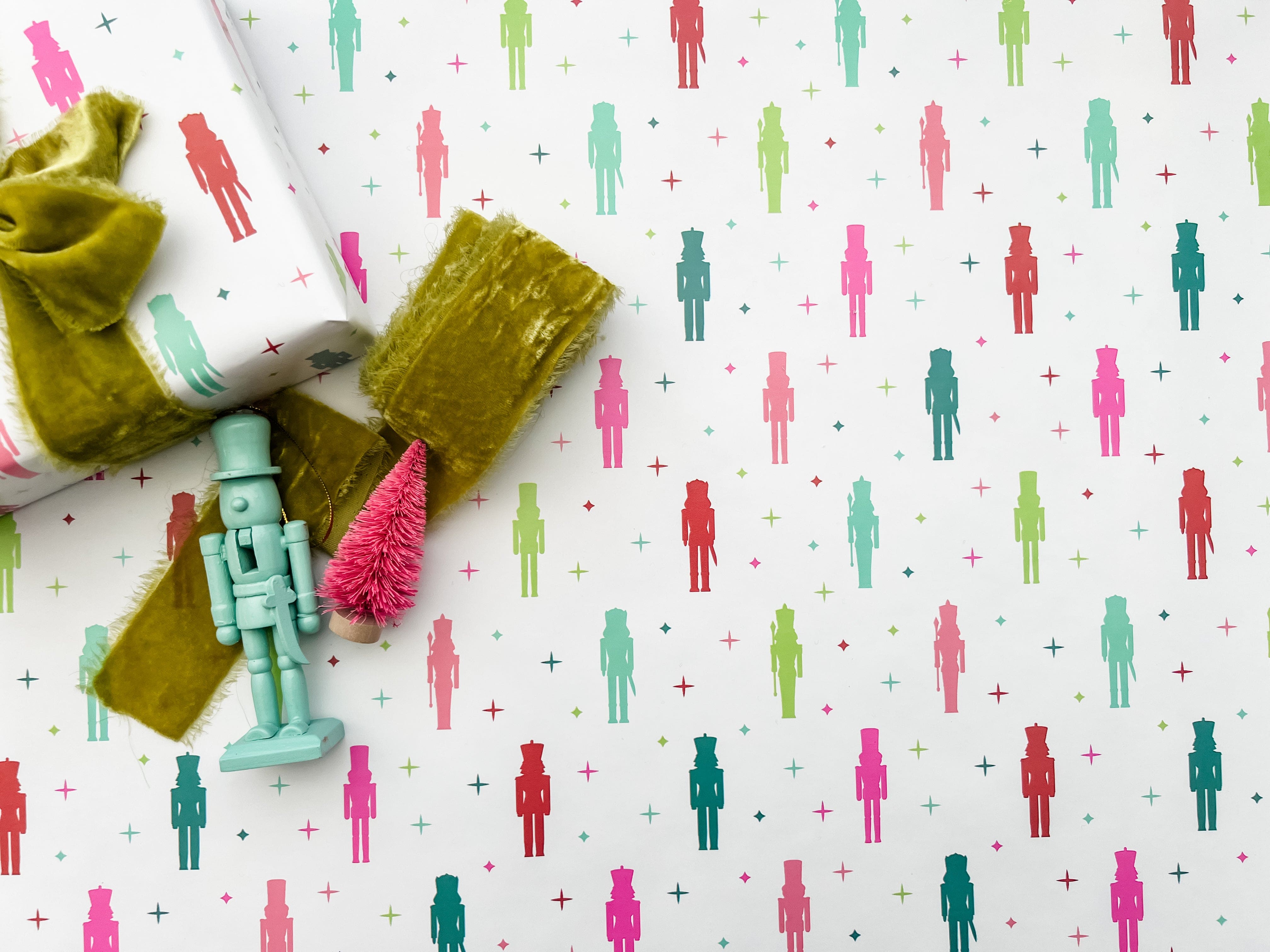 A laid out roll of wrapping paper with a colorful nutcracker pattern featuring nutcracker figures, displayed alongside a green ribbon and a decorative nutcracker.