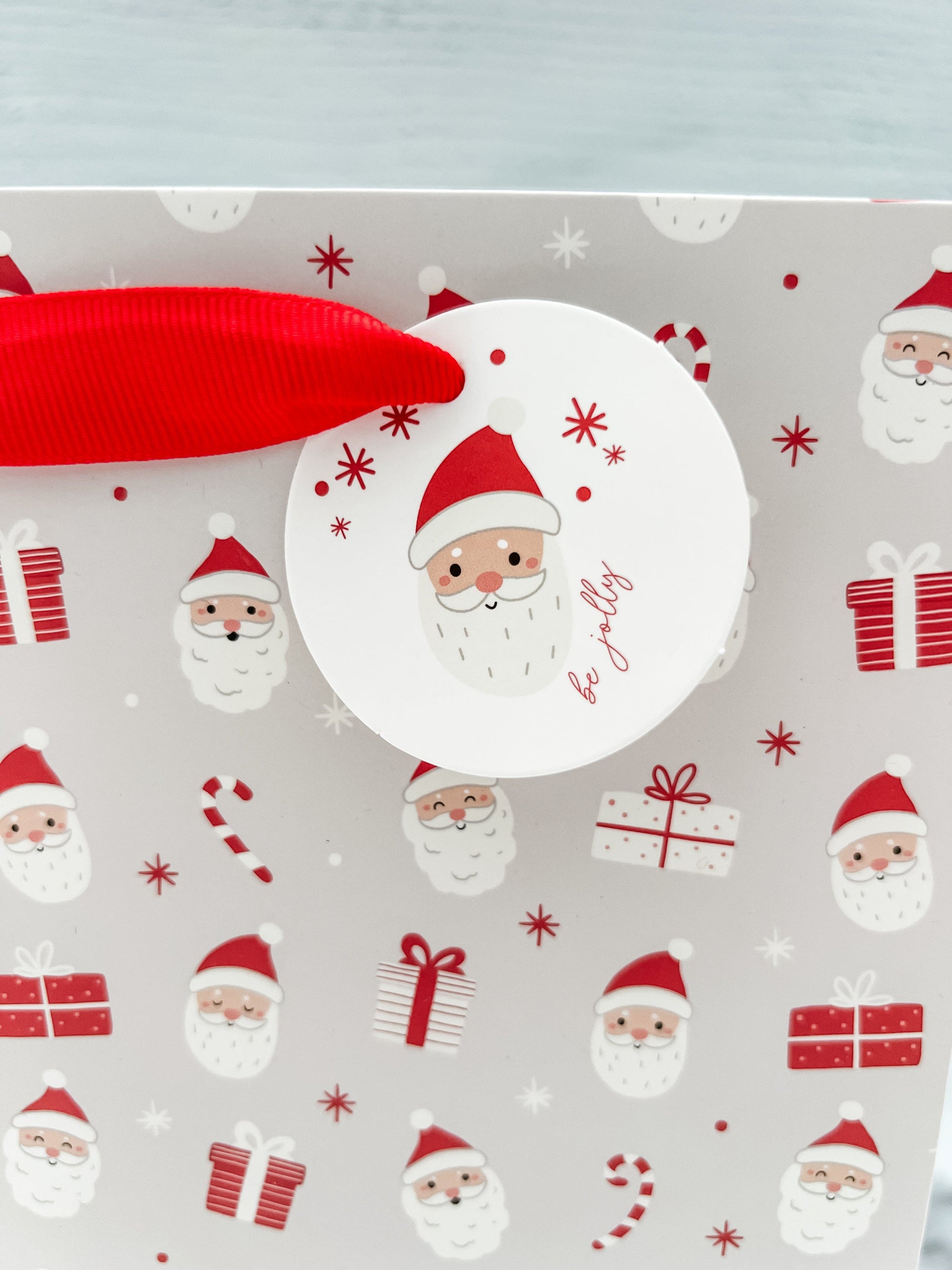 A festive Santa Claus design gift bag with a red ribbon handle and a round tag on the top right corner, featuring a pattern of Santa Claus.
