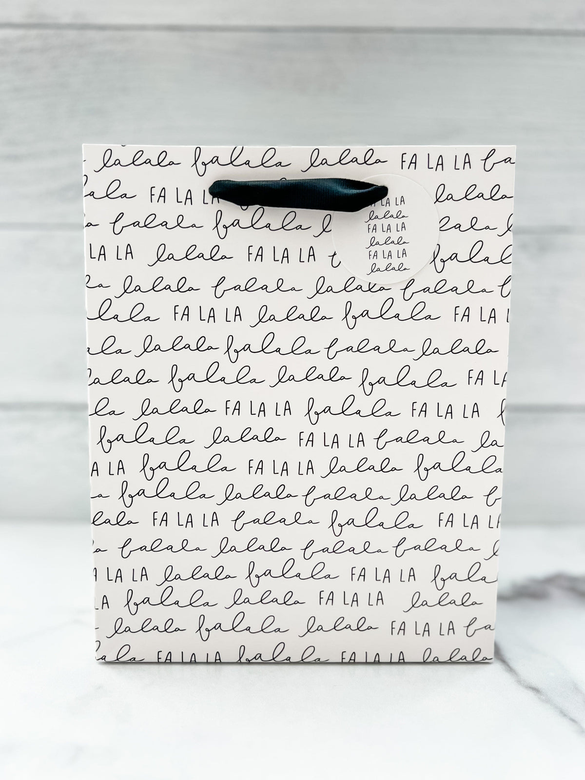 A festive 'Fa La La' text design gift bag placed against a wooden background.