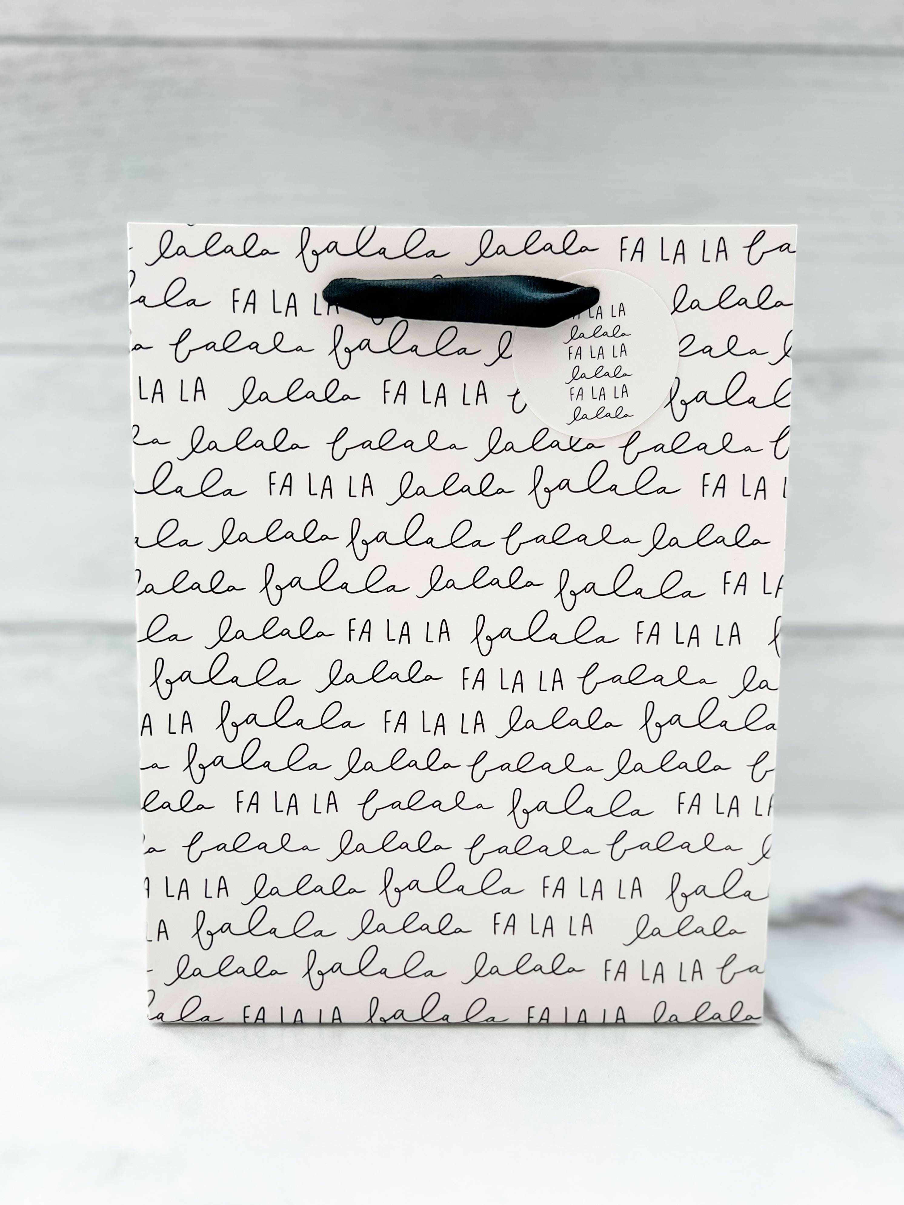 A festive 'Fa La La' text design gift bag placed against a wooden background.