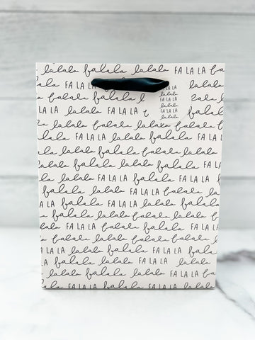 A festive 'Fa La La' text design gift bag placed against a wooden background.