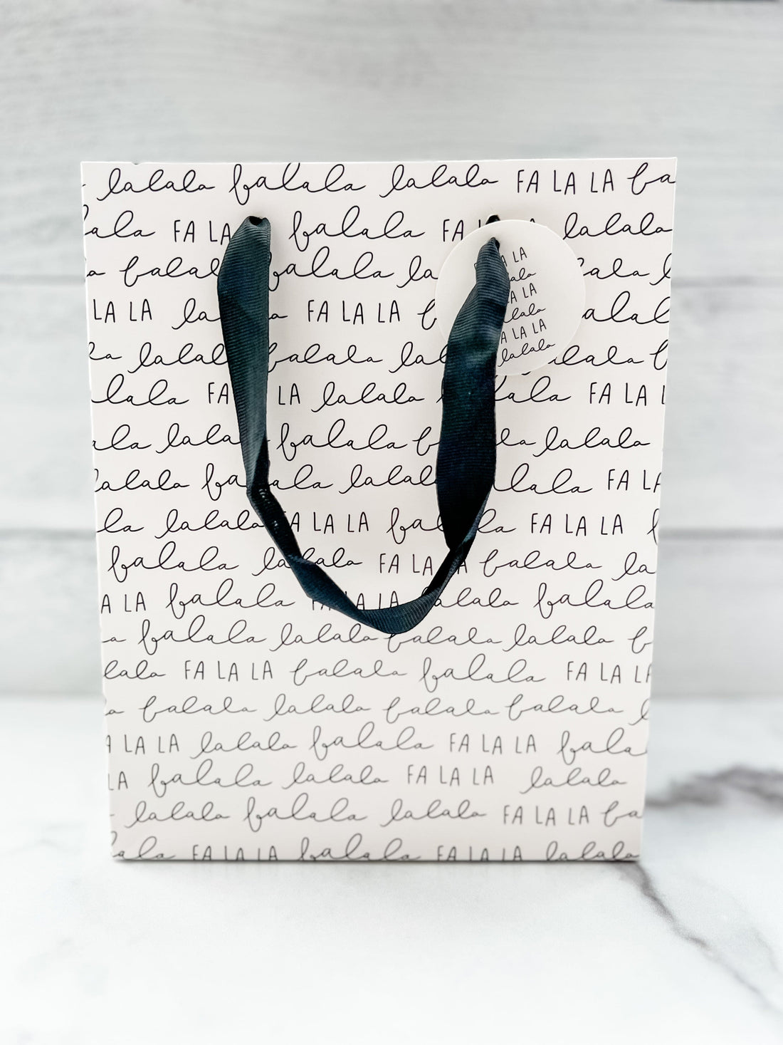A festive 'Fa La La' text design gift bag placed against a wooden background.