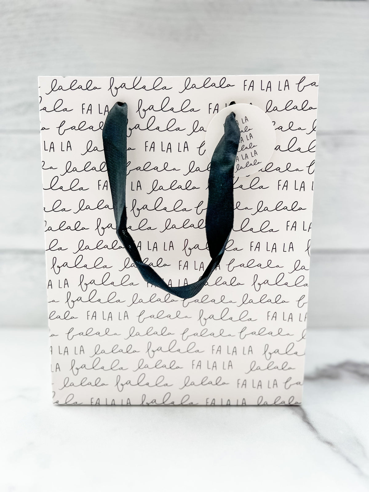 A festive 'Fa La La' text design gift bag placed against a wooden background.