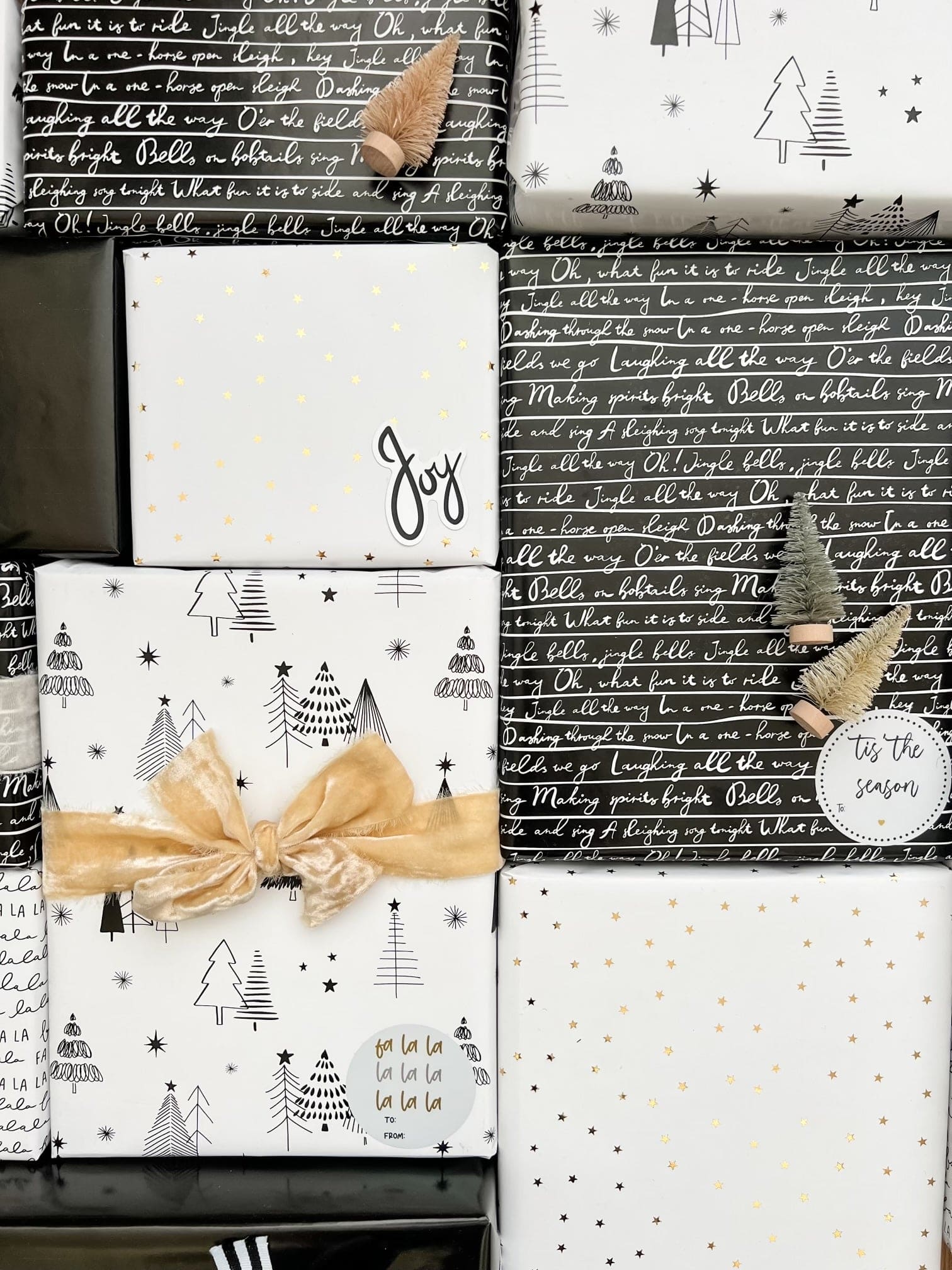 A styled flat-lay of wrapped Christmas gift boxes and sheets of wrapping paper. The designs feature black and white holiday motifs including Christmas trees, stars, and script text, accented with gold foil stars and a gold ribbon bow.