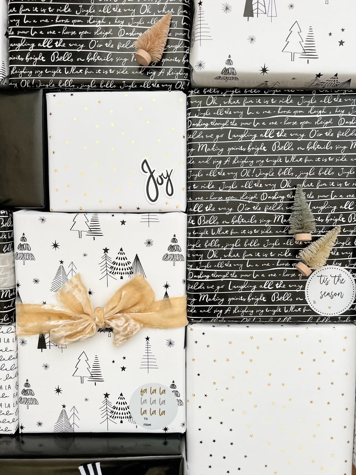 A styled flat-lay of wrapped Christmas gift boxes and sheets of wrapping paper. The designs feature black and white holiday motifs including Christmas trees, stars, and script text, accented with gold foil stars and a gold ribbon bow.