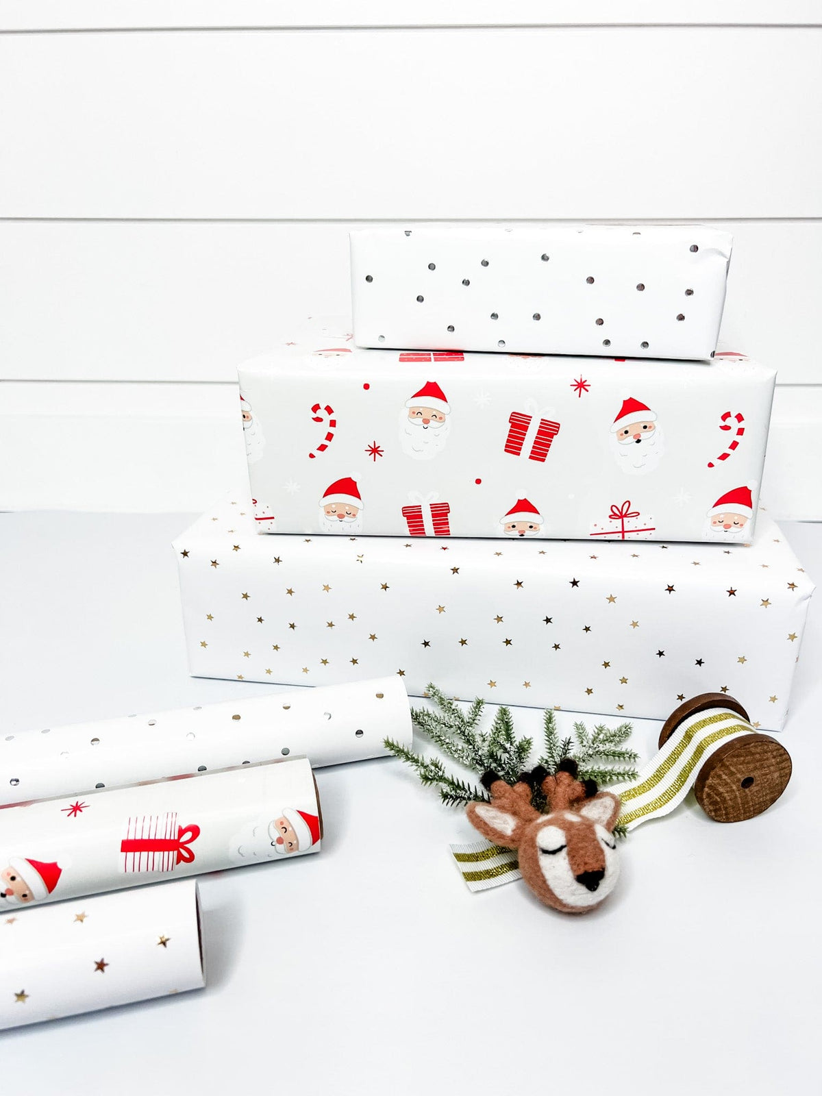 Three rolls of wrapping paper laid out next to each other, with one roll featuring a santa pattern, another with gold stars, and the last one with metallic silver dots. next to a stack of presents