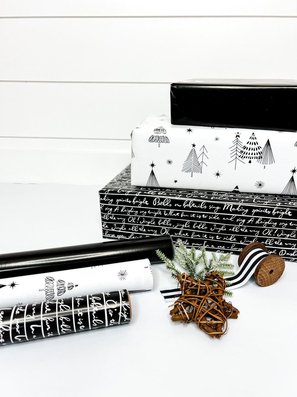 Three rolls of wrapping paper laid out next to each other, with one roll featuring a jingle bells pattern, another with tree silhouettes, and the last one solid black. Presentation decorations like pine cones are also visible.
