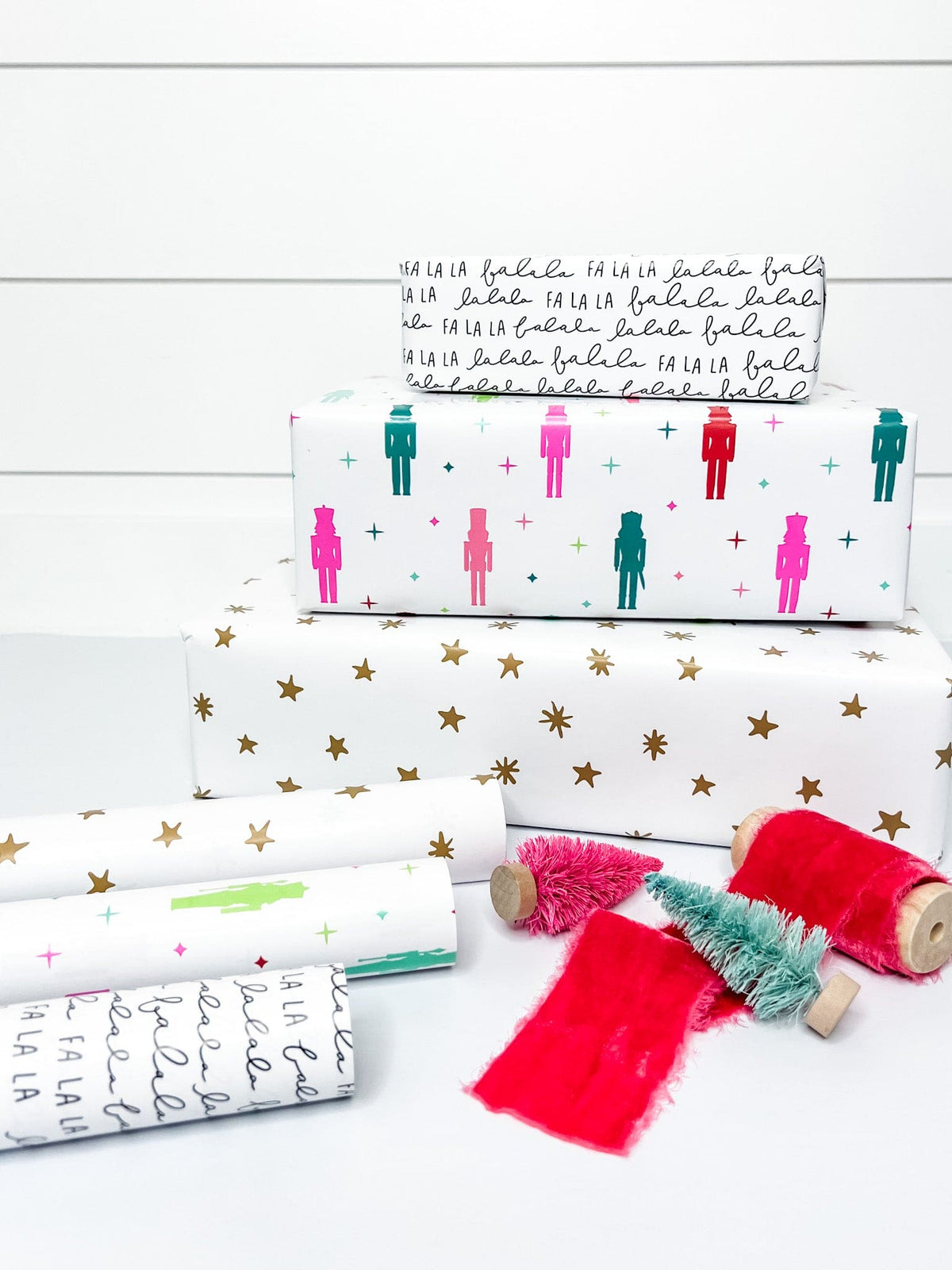 Three rolls of holiday wrapping paper with one featuring colorful nutcracker figures, another with a 'FA LA LA' text and stars, and the last with a gold star pattern. 