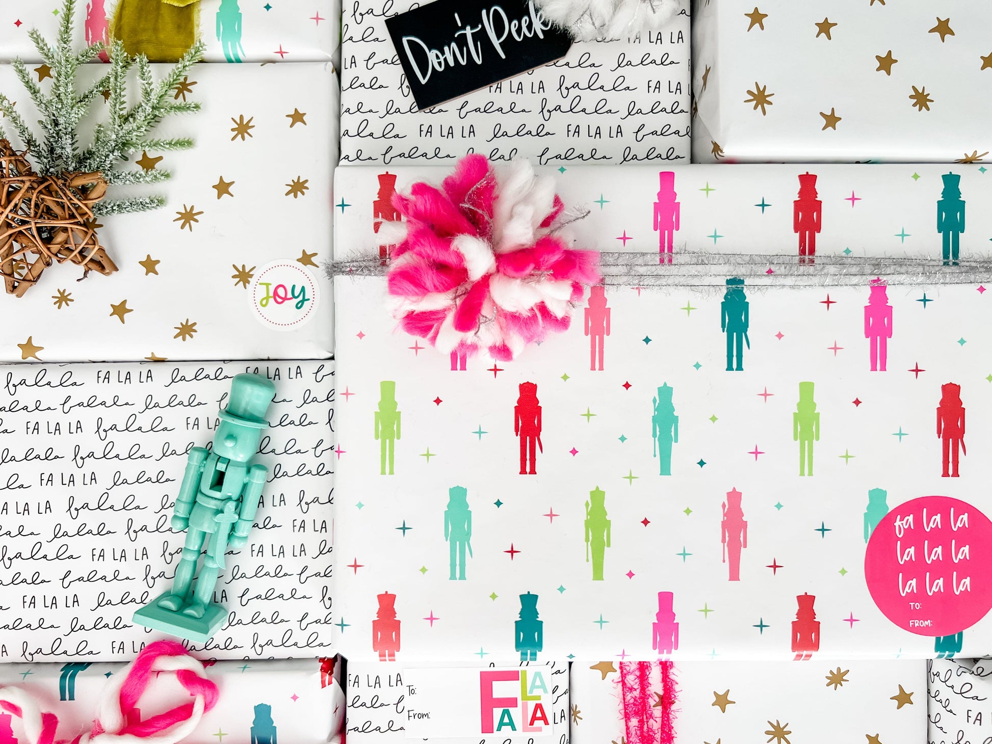 A styled flat-lay of multiple wrapped gift boxes featuring a festive holiday pattern with the words 'Fa La La', colorful silhouettes of figures, gold stars, and a pink pom-pom decoration.